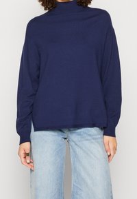 Kvinna iklädd en marinblå polotröja med långa ärmar, kombinerad med ljusblå vida jeans, stående mot en enfärgad bakgrund.