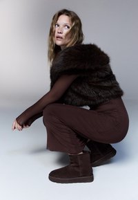 Hnědý krátký vest s kožešinovým vzhledem, dlouhé hnědé rukávy, hnědé upnuté kalhoty a tmavě hnědé kotníkové boty. Modelka squatuje na světlém pozadí.