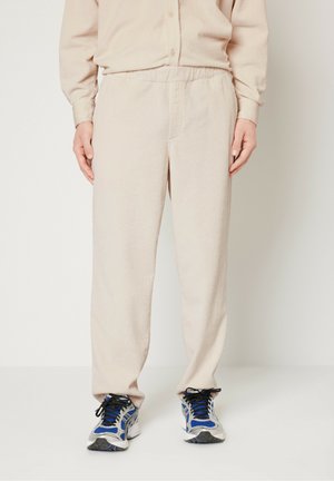 Light beige corduroy pants with elastic waistband, featuring textured stripes. Paired with gray and blue athletic shoes.