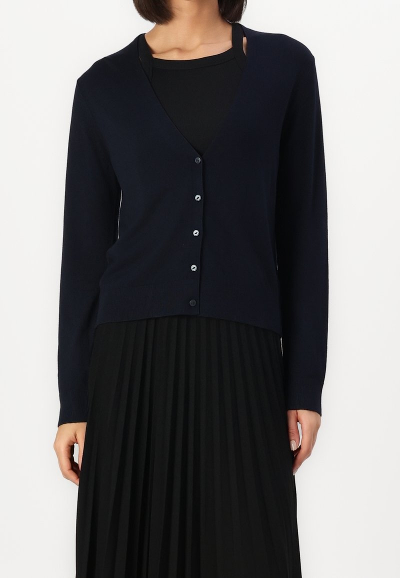 Femme portant un cardigan noir boutonné sur un top noir et une jupe noire plissée, montrée des épaules jusqu'au milieu des cuisses.