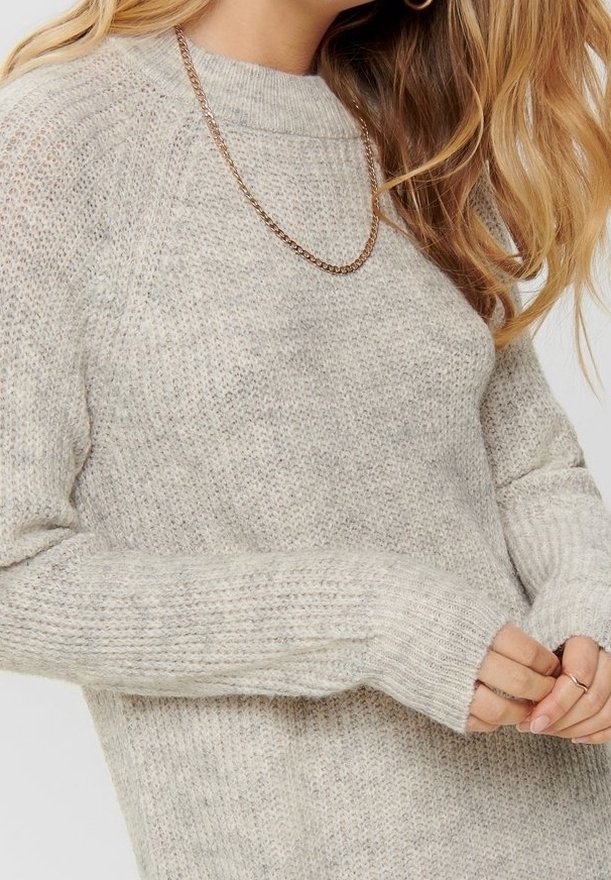 Pull en maille beige clair avec une texture côtelée, manches raglan et col haut. Présente un délicat collier chaîne en or.