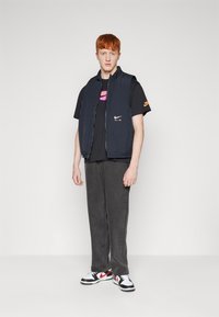 Ναυτικό αμάνικο γιλέκο πάνω από μαύρο μπλουζάκι με μοτίβο σε μωβ και ροζ. Γκρι παντελόνι και μαύρα, άσπρα και κόκκινα αθλητικά παπούτσια ολοκληρώνουν την εμφάνιση.