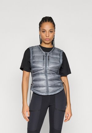 Gilet matelassé gris avec fermeture éclair, présentant un col haut et des côtés ajustables. Porté sur une chemise noire, associé à un leggings sombre.