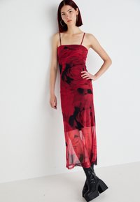 Vestido maxi de seda vermelha com um padrão floral, apresentando alças finas e um corpete ajustado, combinado com botas pretas de salto robusto.