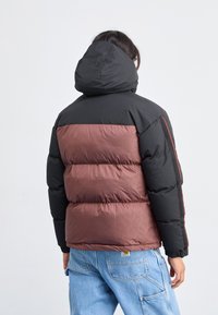 Chaqueta acolchada con parte superior negra y parte inferior marrón, con una textura acolchada. Incluye capucha y un sutil patrón en la tela marrón.