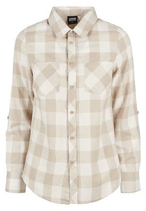 Person iført beige ternet skjorte med knapper og opkrøllede ærmer, lyseblå jeans og sorte sko står på en grå gulvbelægning mod en ensfarvet baggrund.