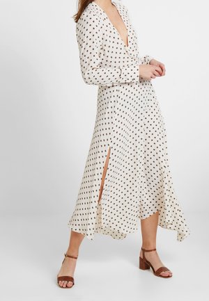 Lange crèmekleurige jurk met bruine stippen, diepe V-hals, lange mouwen en zijsplitten. Draagt het met bruine hoge sandaaltjes. Glad fabrieksweefsel.