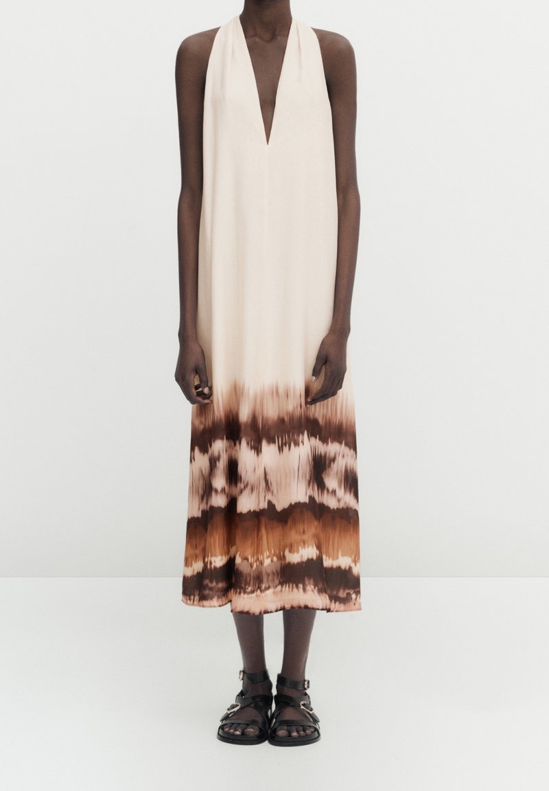 Robe dos nu crème avec un motif tie-dye, passant du brun clair au brun foncé à l'ourlet ; associée à des sandales noires à brides.
