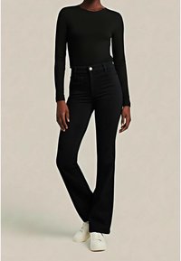 Personne portant un haut noir à manches longues et un pantalon noir évasé taille haute, debout avec un genou légèrement plié et des chaussures blanches.