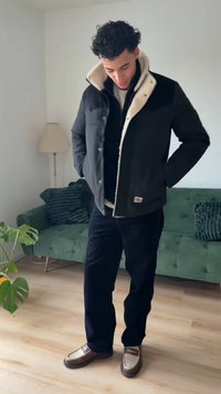 Black outer shell jacket with white fleece lining, paired with black pants and two-tone loafers, set against a green couch backdrop.