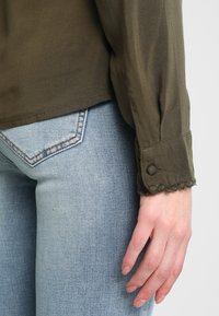 Olivgrön blus med långa ärmar, rundad krage och dekorativa scalloped manschetter, kombinerad med ljusblå denimjeans med bakfickor.