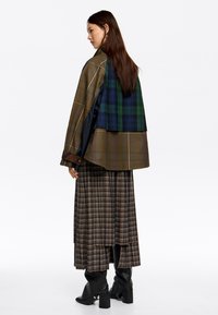 Prevelik plašč s karirnim vzorcem v zemeljskih tonih, obložen s kontrastnim zelenim in modrim karirastim vzorcem; v kombinaciji z dolgim karirastim krilom in črnimi čevlji.