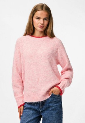 Kvinde med langt brunt hår, iført en blød lyserød sweater med rød kant ved kraven og håndleddene, parret med blå jeans, og står med hånden i lommen.