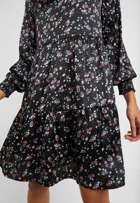 Robe noire fleurie à manches longues, taille froncée et ourlet à volants, portée par une personne avec une main visible à côté.