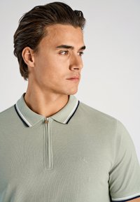 Koszulka polo z jasnej zielonej tkaniny z zamkiem błyskawicznym, z kołnierzykiem w granatowo-białe paski i subtelnym logo.