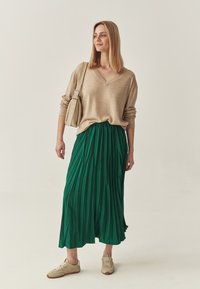 Suéter de viscosa beige claro con escote en V combinado con una larga falda verde plisada. El conjunto se completa con zapatillas beige y un bolso de hombro beige claro.