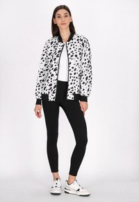 Zwart-wit gespikkelde bomberjack met zwarte accenten, gecombineerd met zwarte leggings en witte sneakers, op een effen achtergrond.