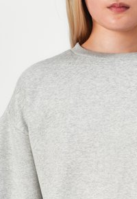Närbild av en person som bär en ljusgrå stickad tröja med rund halsringning, som visar läppar, nacke och övre bröst mot en helt vit bakgrund.