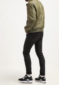 Olivgrüne Pufferjacke mit strukturiertem Material, schwarze Skinny Jeans und schwarze Sneakers mit weißen Akzenten und Logo.
