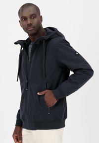Uomo che indossa una giacca con cappuccio blu navy dotata di lacci e tasche con zip, in piedi con una mano in tasca su uno sfondo bianco.