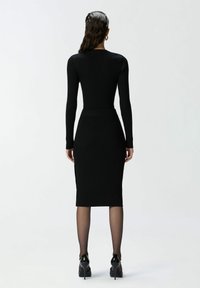Frau mit langen dunklen Haaren, die ein enganliegendes schwarzes Rippkleid, schwarze, durchsichtige Strumpfhosen und schwarze High Heels trägt, und mit dem Rücken zur Kamera vor einem weißen Hintergrund steht.