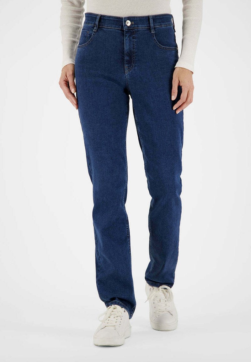 Personne portant un jean bleu foncé à jambes droites et des baskets blanches, debout devant un fond blanc uni.