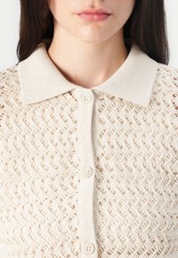 Lichtbeige gebreide cardigan met een gestructureerd, open patroon. Heeft een contrasterende platte kraag en vier kleine, ronde knopen aan de voorkant.