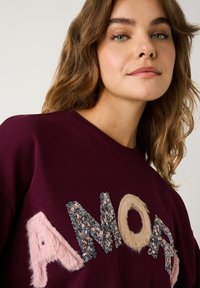 Eine Frau mit hellbraunem, welligem Haar trägt einen burgunderfarbenen Pullover mit strukturierten Buchstaben "AMOR" in Pink, Beige und mit Paillettenverzierungen.