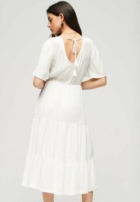 Vestido blanco con mangas cortas, falda en capas y espalda descubierta sujeta por un lazo. Presenta un delicado acabado en el escote y las mangas.