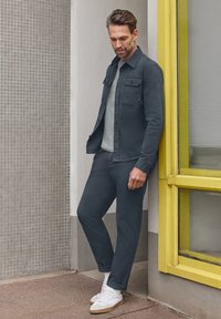 Mężczyzna w ciemnej kurtce i spodniach, białe trampki, opierający się o okno z żółtą ramą na ścianie z płytek, patrzący w dół.