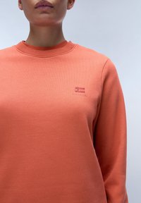 Oranje sweatshirt met een geribde halslijn en lange mouwen. Bevat een klein geborduurd logo op de linkerborst. Zachte textuur.