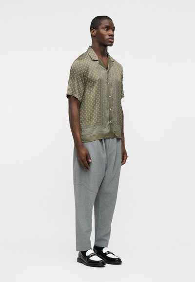 Άνδρας που στέκεται φορώντας πουκάμισο με κοντά μανίκια και μοτίβο, φαρδιά γκρι παντελόνια, μαύρα και άσπρα μοκασίνια, μπροστά σε απλό ανοιχτό φόντο.