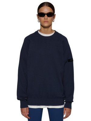 Kvinde iført mørkeblå crewneck sweater over hvid skjorte, blå bukser, sorte slanke solbriller og sølv hoop øreringe, står foran hvid baggrund.