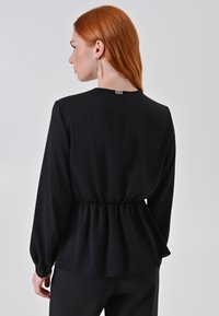 Blusa nera con maniche lunghe, vita arricciata, texture liscia e scollo rotondo con un piccolo logo sul retro.