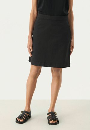 Kvinde iført sort knælængde nederdel, sort ærmeløs top og sorte flettede sandaler, stående mod en ensfarvet baggrund.