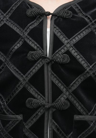 Μαύρο σακάκι από βελούδο με σχεδιασμό καπιτονέ, δεμένα τελειώματα και μπροστινές κλειδαριές με κόμπο. Περιλαμβάνει δύο τσέπες και περίτεχνες λεπτομέρειες ραφής.