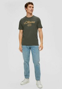 Olive green short-sleeve t-shirt featuring white and orange text. Paired with light blue denim jeans and white sneakers.