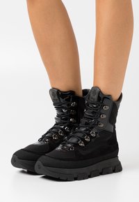 Scarpe da trekking nere caratterizzate da una combinazione di materiale verniciato e scamosciato, occhielli in metallo, suola in gomma spessa e design con lacci.