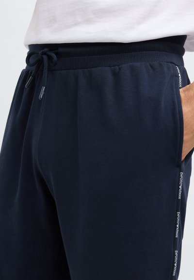 Tengerészkék színű, puha anyagból készült melegítőnadrág, elasztikus derékrésszel, húzózsinór részlettel és oldalzsebekkel, melyeken márkázott díszítések találhatók.