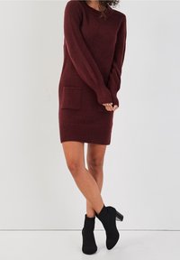 Robe pull en tricot bordeaux à manches longues, avec deux poches frontales, des poignets côtelés et un ourlet côtelé. Associée à des bottines noires.