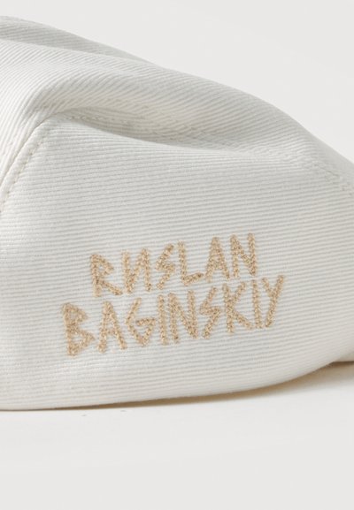 Κοντινή φωτογραφία λευκού υφασμάτινου υφάσματος με μπεζ κεντημένο κείμενο "RUSLAN BAGINSKIY" στο πλάι ενός καπέλου ή μπερέ.
