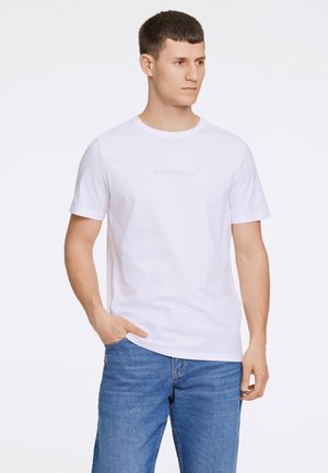 Vit bomull t-shirt med rund hals och korta ärmar; har subtilt tryckt text "LINDBERGH" på framsidan. Matchas med blå jeans.