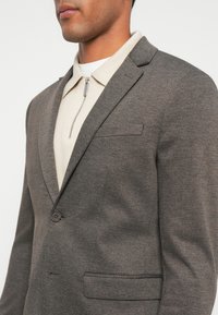 Hnedý textúrovaný blazer s klopami, jedným gombíkom na zapínanie a jednou náprsníkovou kapsou. Nosí sa cez svetlú košeľu so zipsom a golierom.
