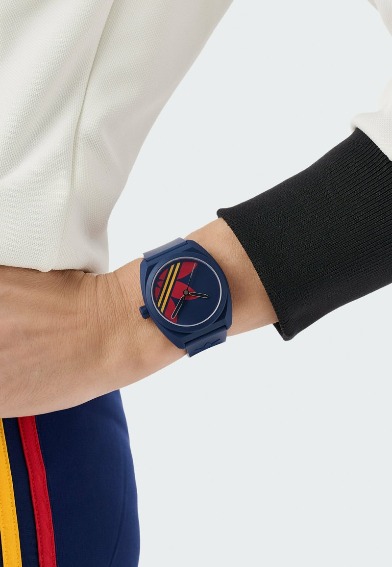 Poignet portant une montre bleu marine avec un design géométrique rouge et jaune, associée à une manche blanche et un pantalon bleu marine avec des rayures rouges et jaunes.