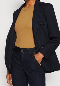 Námořnická sako s upraveným střihem, čtyřmi knoflíky a kapsami, nošené přes béžové proužkované tričko. Tmavé džíny s detaily zipovaných kapes.