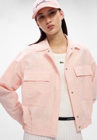 Veste rayée rose avec deux poches avant, fermeture à bouton-pression et un tissu texturé. Portée par-dessus un haut blanc avec un détail de taille noire.