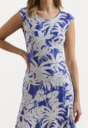 Femme portant une robe sans manches avec un motif de feuilles tropicales bleu et blanc, main posée sur la hanche.