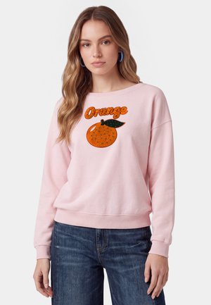 Giovane donna che indossa una felpa rosa chiaro con un frutto arancione di perline e la parola "Orange" in perline arancioni sul petto.