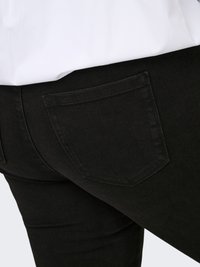 Juodi džinsai iš džinso audinio su matomomis siūlėmis ant užpakalinės kišenės, dėvimi su įkišta balta palaidine, akcentuojant juosmenį ir viršutinę šlaunies dalį.