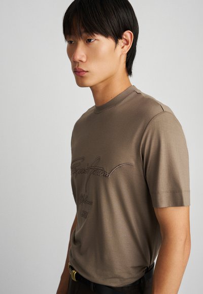 Braunes T-Shirt mit kurzen Ärmeln und glatter Textur, mit gesticktem Schriftzug auf der Vorderseite. Der Kragen ist rund und die Passform ist normal.